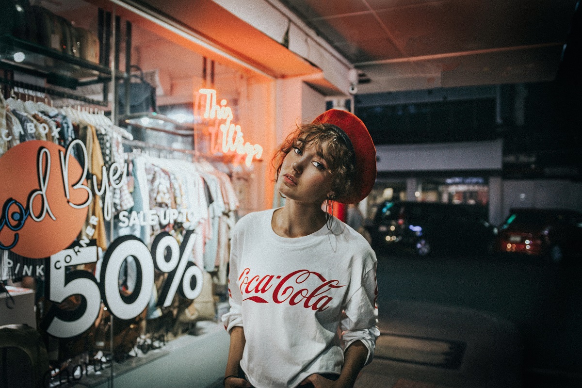 Mulher jovem com boina vermelha, camisa branca com o logo vermelho da Coca-Cola em frente a uma loja onde se lê 50%
